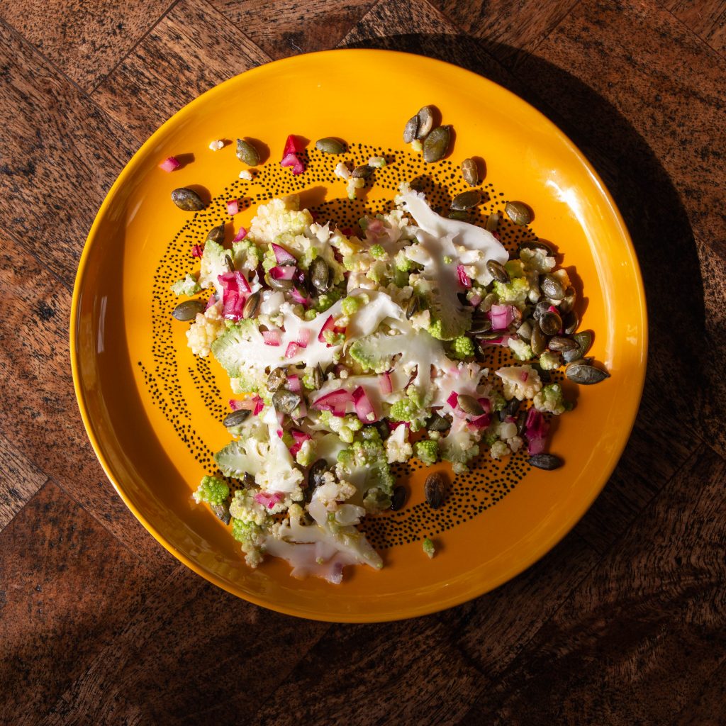 Image features a cauliflower and red onion pickle salad topped with sunflower seeds on a yellow plate with a black pattern on a dark wooden table.