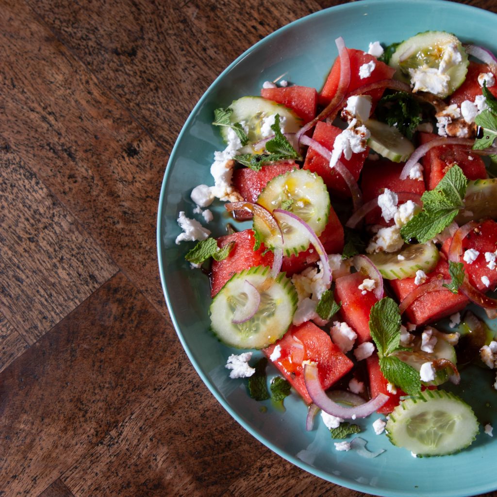 Image features a watermelon and cucumber salad on a blue plate with a wooden background