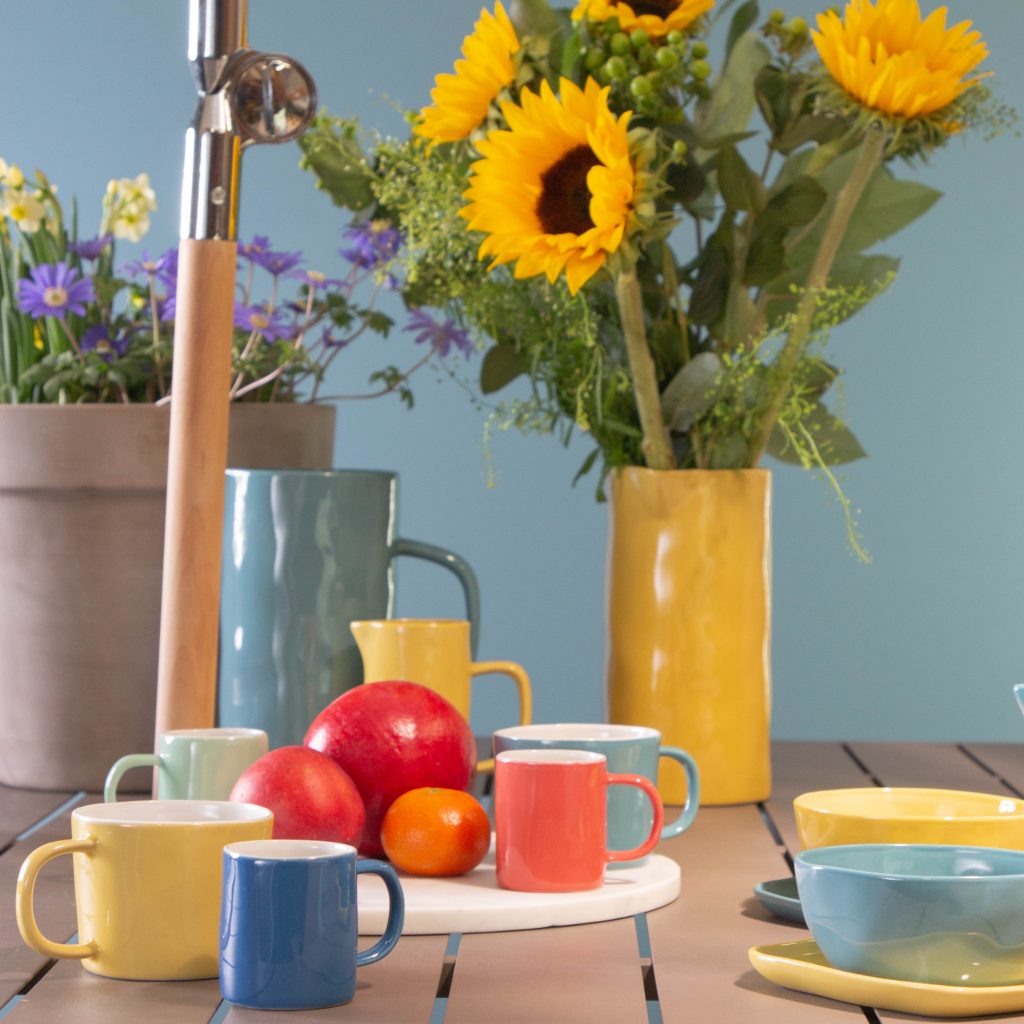 Images features a colourful tableware set with sunflowers in the background and piles of fruit