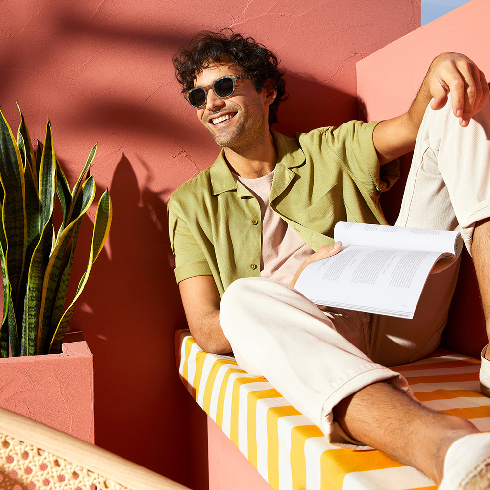 Image features a man wearing Izipizi sunglasses outside while reading a book against a colourful pink and yellow background