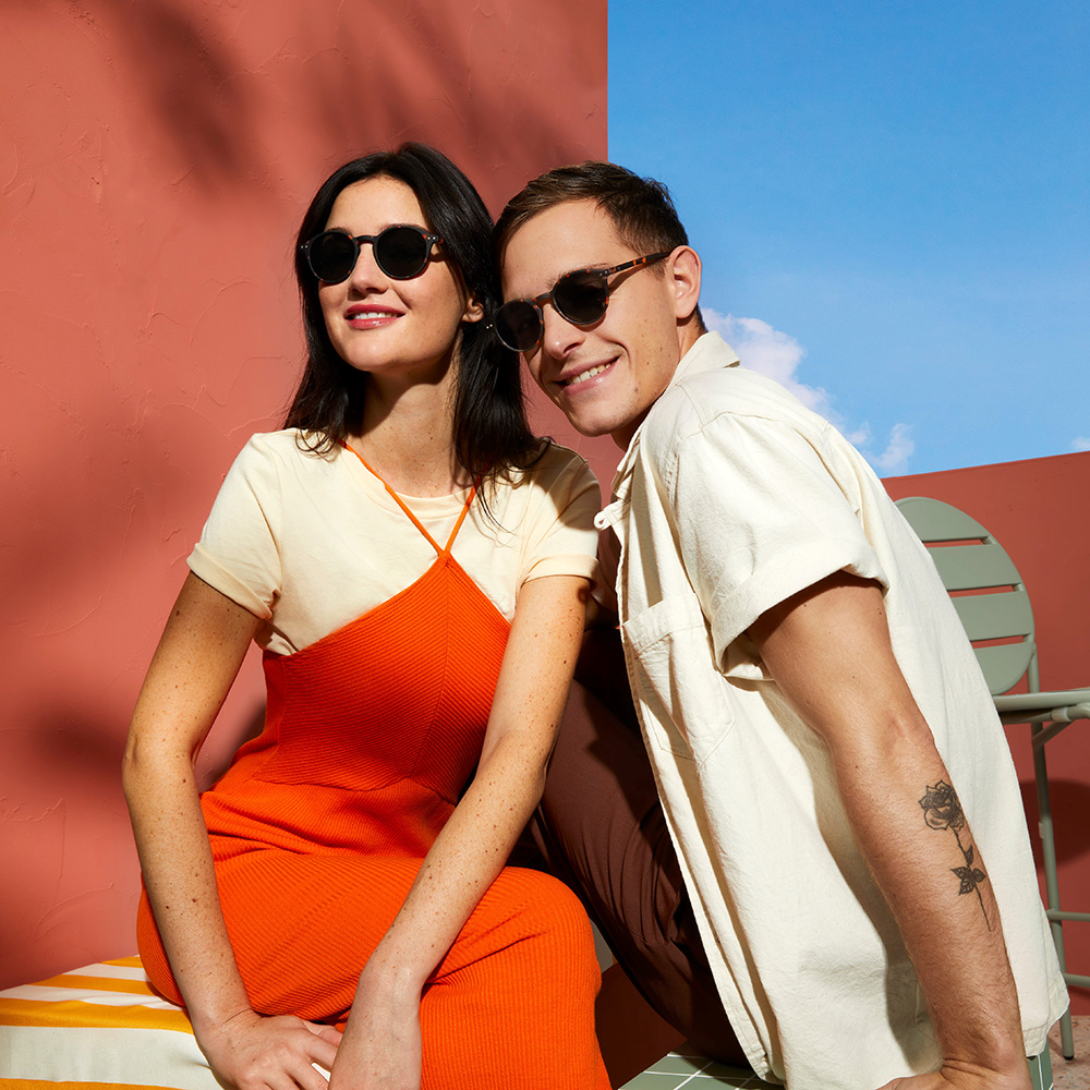 Image features two people against a red background with the sky in the top corner. They are wearing pairs of Izipizi sunglasses