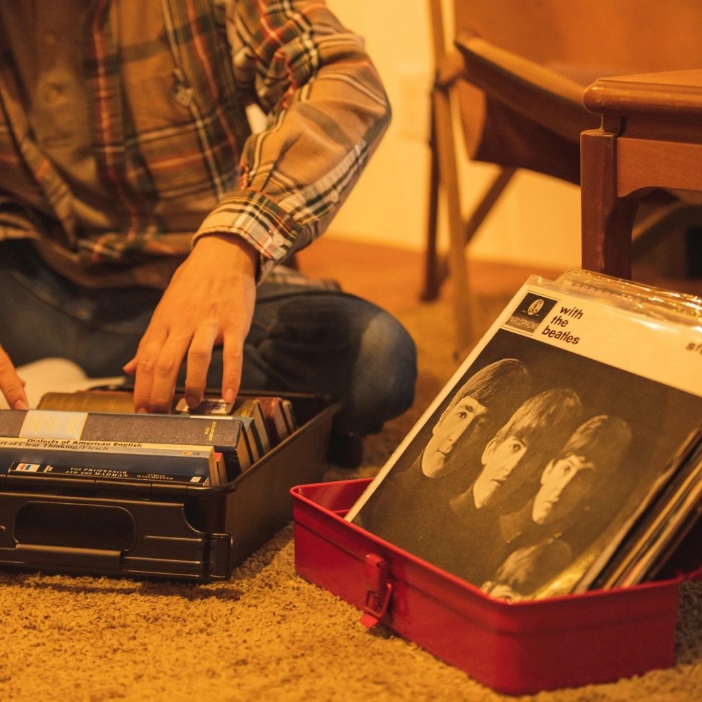 Image features a red tool box holding records with a man sitting next to it flicking through another box of casettes